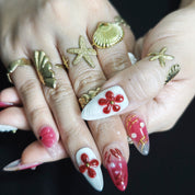 White and red almond press-on nails with 3D floral designs, rhinestones, pearls, and gold bead accents for a luxurious handmade look.